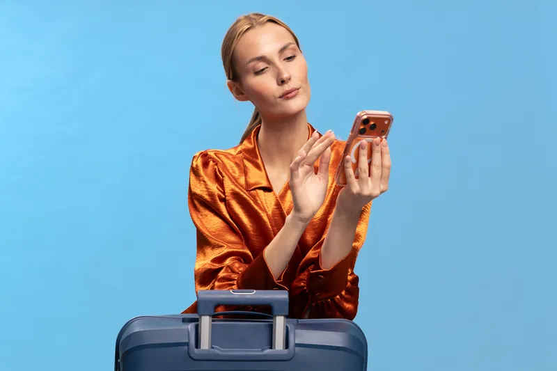 Young woman in shiny orange blouse checking smartphone while standing behind blue suitcase on light blue background. Concept of travel planning, lifestyle, and modern communication.