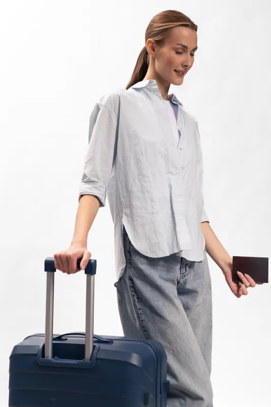 Young woman in casual clothes holding a passport and pulling a suitcase. She is ready for travel, standing against a white background with a relaxed and confident expression.