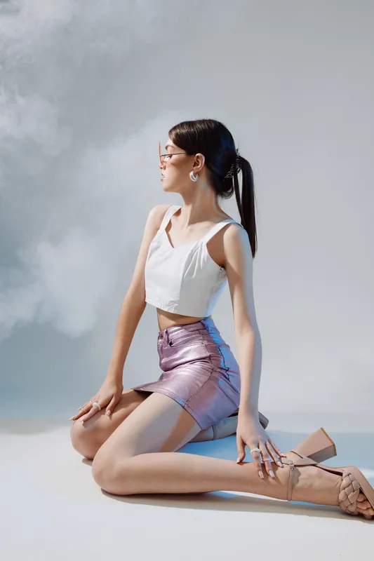 Young woman beauty fashion model kneeling in a shiny skirt and white top. She poses confidently in studio lighting with a soft gray background. Concept of style and elegance.