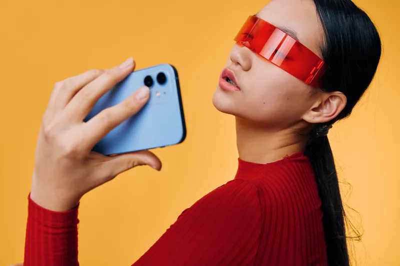 Woman in red sweater and futuristic red glasses holds smartphone at face level against yellow background for digital communication and technology concept in modern lifestyle.