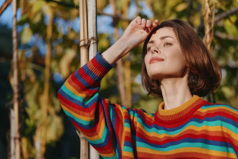 Woman in rainbow striped sweater with short brown hair, smiling and leaning on a bamboo pole in sunny outdoor tropical portrait. Sunlight, relaxed travel lifestyle fashion mood.