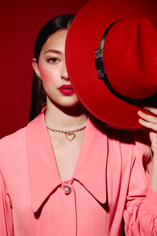 Young woman with glossy red lipstick and pink blush wears a pink blazer and a pearl necklace while partially hiding her face behind a red wide-brimmed hat in studio lighting.