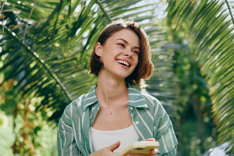 Bright outdoor portrait of a cheerful woman holding a smartphone, surrounded by lush tropical plants.