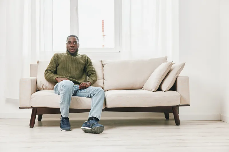 A relaxed African American man sitting on a modern beige sofa, showcasing a casual style and a calm expression in a bright, minimalistic living room
