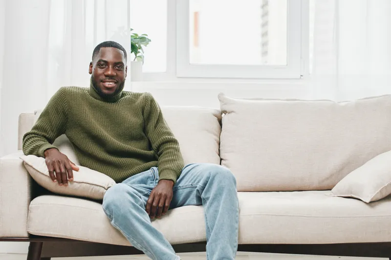 Smiling young Black man in a cozy green sweater sitting on a beige sofa, embodying relaxation and comfort in a bright indoor space filled with natural light
