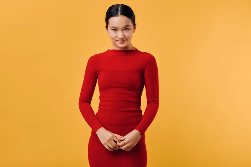 Young woman in red dress standing against orange background with clasped hands, confident expression, and relaxed posture in studio portrait with simple colorful backdrop.