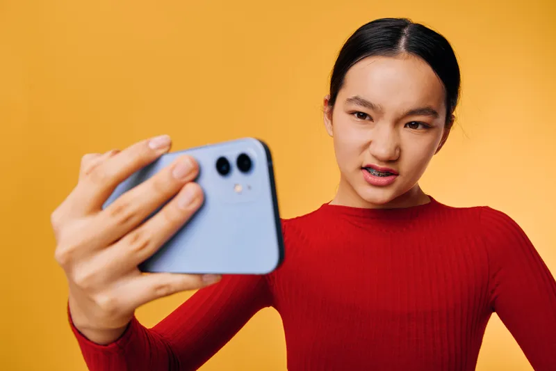 Young woman in red sweater making frustrated face while holding smartphone close for selfie or video call on bright orange background with casual expression and focused gaze.
