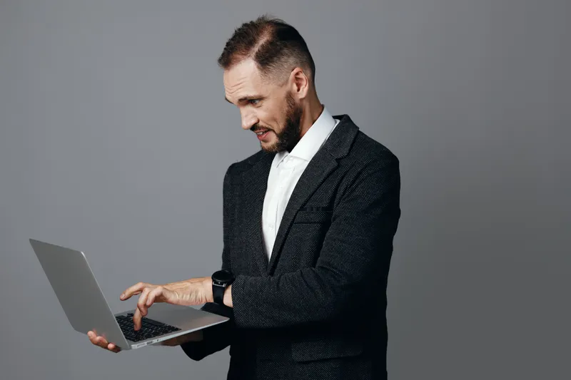 A professional man in a tailored suit uses a laptop, displaying confident focus and calm determination on a solid colored background, isolated from any distractions.