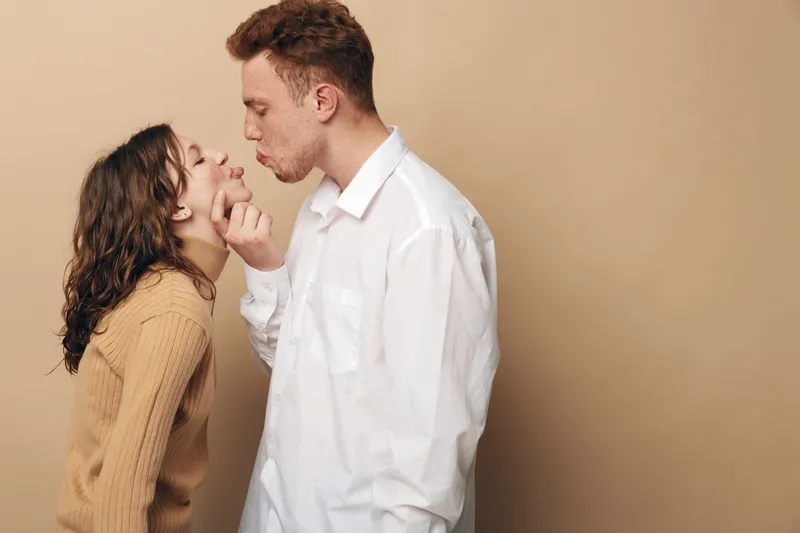 Young couple in casual clothes shares a tender moment, man gently holding woman’s chin while leaning in for a kiss. Beige background creates warm and intimate atmosphere of love and connection.