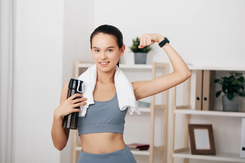 A fit woman in sportswear flexes her bicep, holds a water bottle and towel, celebrating strength during a home workout in a bright, minimal room.