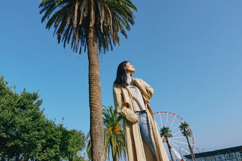 Palm trees frame a stylishly dressed woman in a long coat under a clear blue sky, park setting with a distant wheel for a bright lifestyle and travel moment