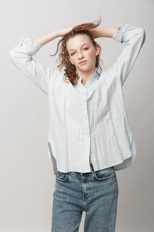 Young woman with curly hair wearing white shirt and blue jeans poses with hands in hair against plain background. Casual style, natural look, and relaxed mood concept.