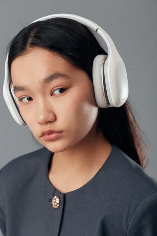 Young woman with long dark hair wearing white headphones and gray top is looking sideways on gray background. Closeup, casual style with natural makeup and thoughtful expression on face.