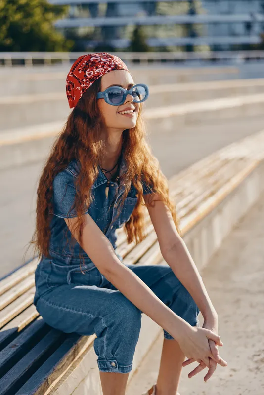 Lifestyle woman in modern boho-western denim street style wearing sunglasses and a red bandana, sitting outdoors with warm film color tones and relaxed mood.
