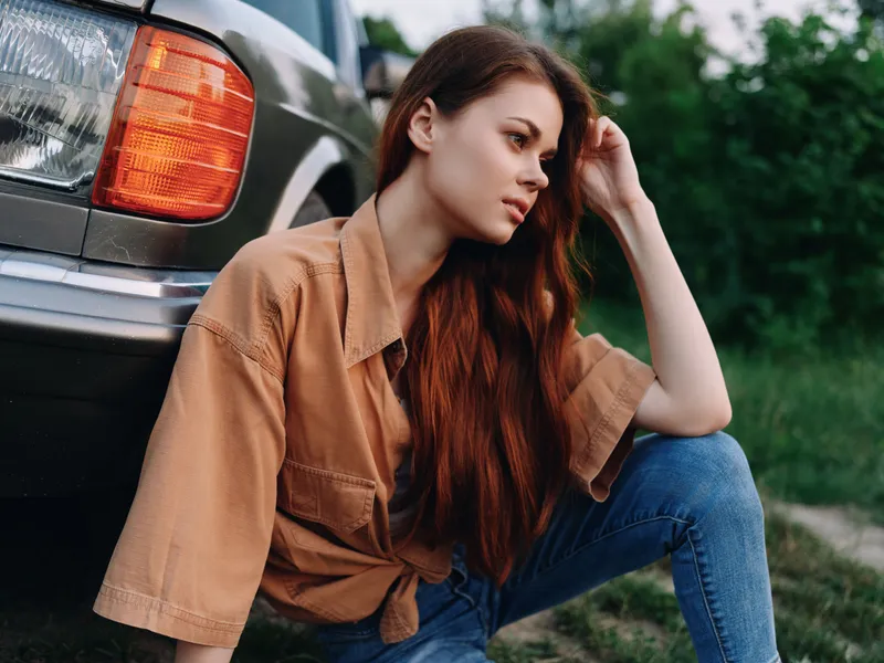 Young woman in casual attire resting by a car, showcasing relaxed emotions and a natural outdoor setting in soft colors.