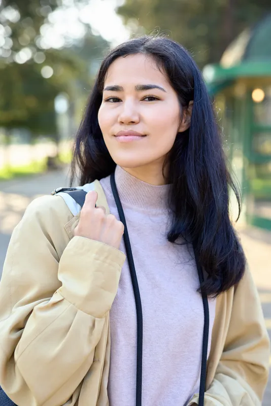 Outdoor woman in a casual jacket and soft sweater with a backpack stands in a sunny park, conveying relaxed style, everyday fashion, and quiet confidence.