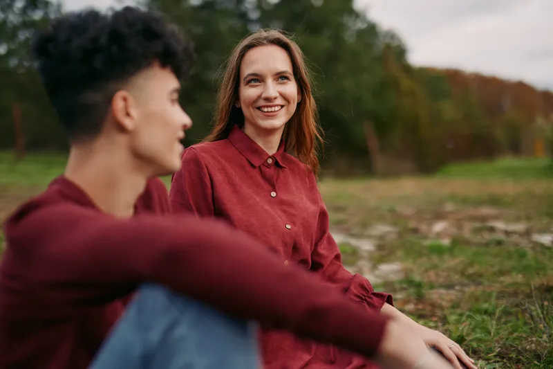 An authentic candid moment shows a smiling woman in a red shirt seated in a grassy field, radiating genuine connection and credibility through a simple, natural pose.