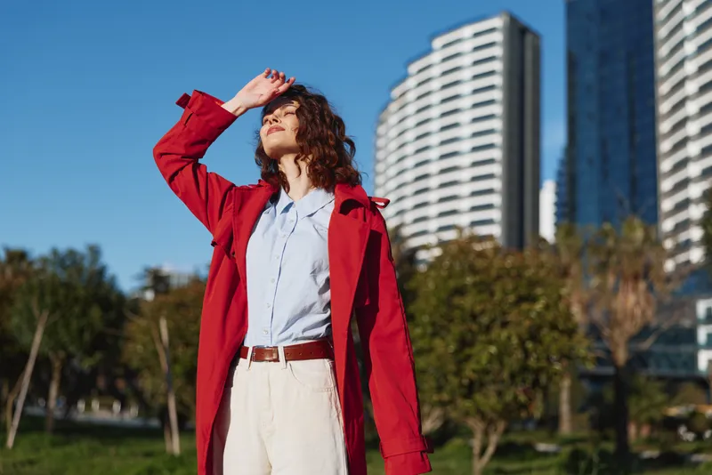 A young woman in a red coat shields her eyes from the sun while posing in a city park with modern highrise buildings behind.