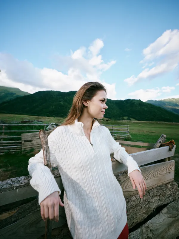Woman in a white knit sweater leaning on a wooden fence in a rural landscape with rolling hills, blue sky and clouds, a relaxed peaceful outdoor moment