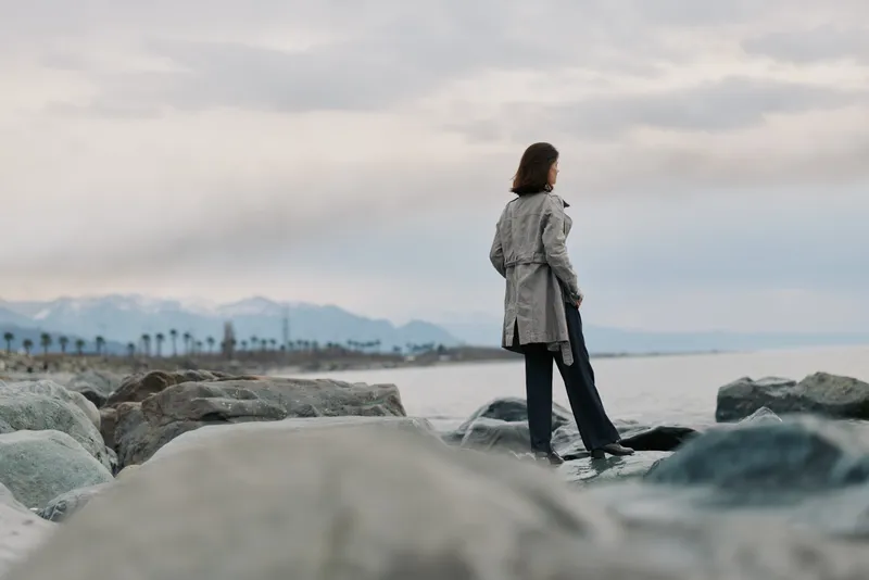 Woman in trench coat stands on rocky shore facing ocean under cloudy sky. Calm coastal landscape with distant mountains and palm trees creates peaceful mood and outdoor concept.