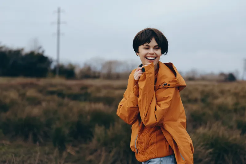 Boy smiling outdoors orange jacket portrait lifestyle nature autumn, young child standing in grass field, orange coat hoodie, relaxed happy face, soft blurred countryside background