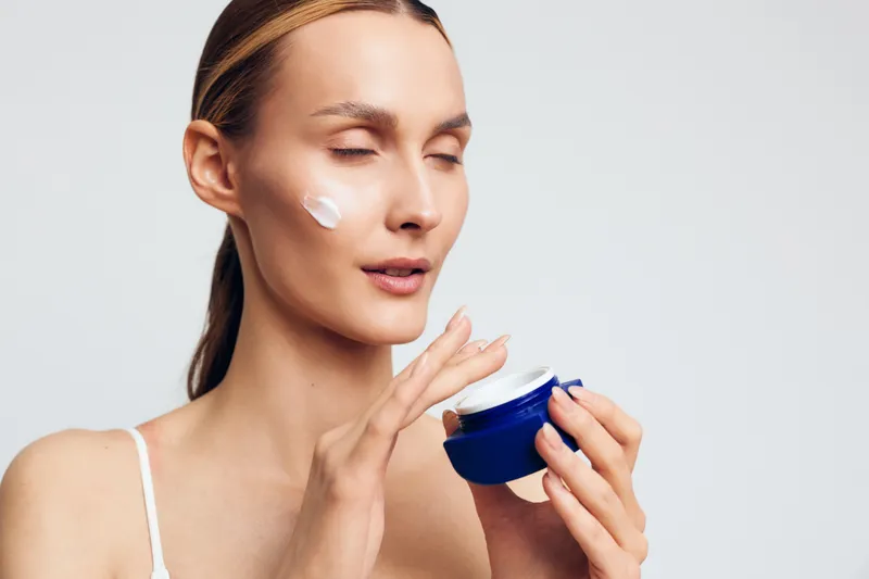 Young woman applying face cream with eyes closed, smooth skin, gentle expression, natural beauty, skincare routine, white background, soft lighting, close-up portrait.