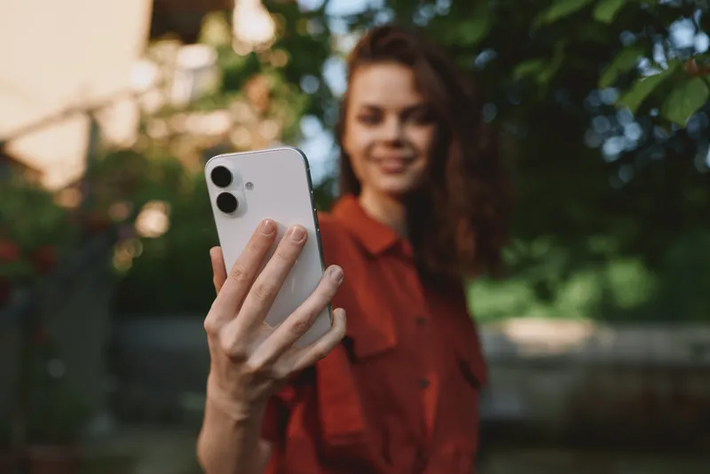 Smiling woman outdoors holding a smartphone for a selfie, casual shirt, blurred greenery in the background, lifestyle moment highlighting connection, social media, and personal expression