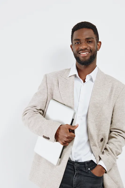 Confident smiling young African American man in beige blazer and white shirt holding a closed laptop, standing against plain light background, casual professional look, studio portrait. People lifesty