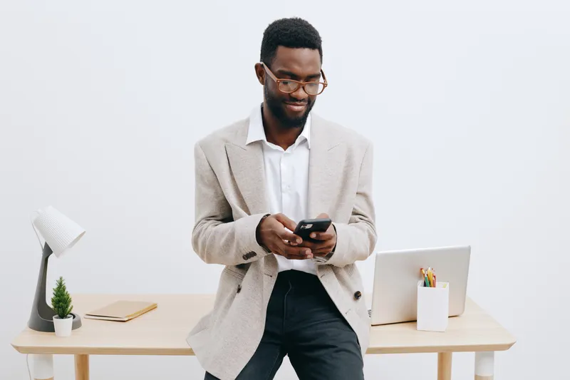 Professional young African American man in smart casual clothing using smartphone in modern office setting with minimalist decor and natural light, representing workplace communication and technology 