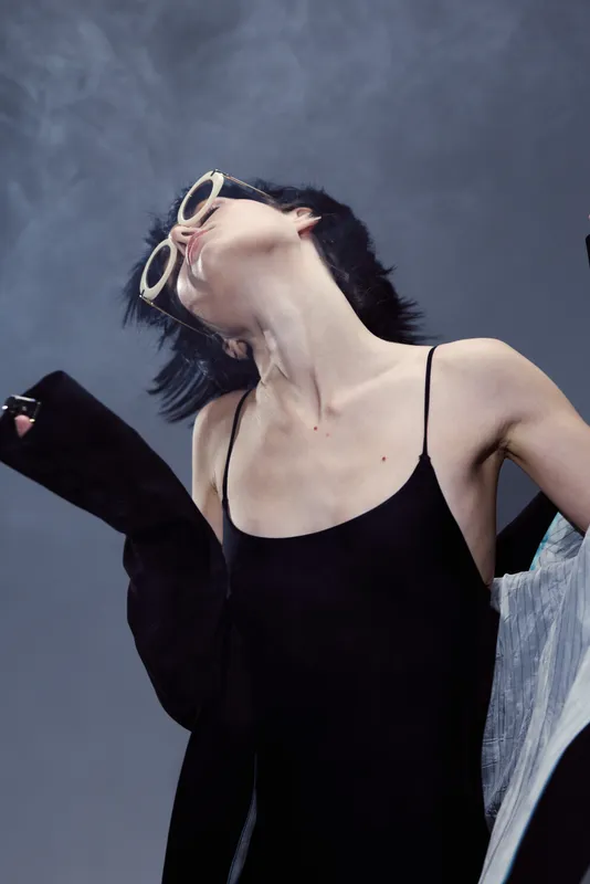 Black-haired woman wearing sunglasses and black dress posing with head tilted back and arms raised in dramatic motion.