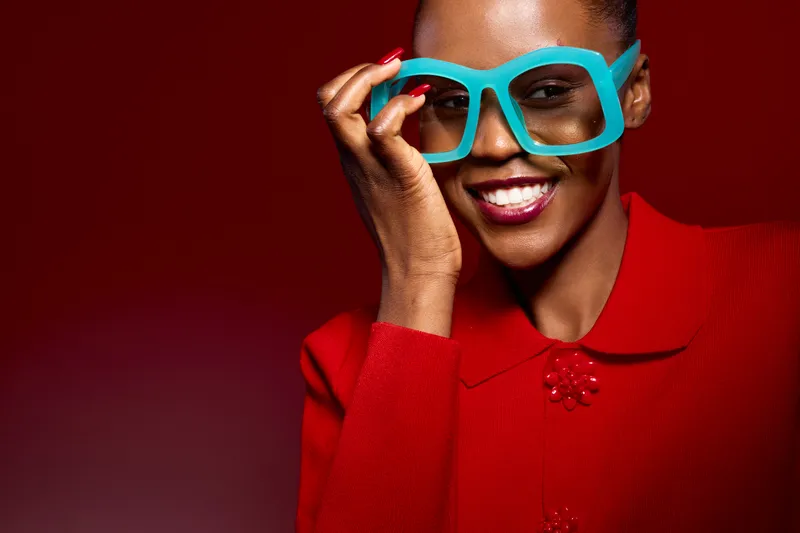 Smiling black woman with dark skin wearing bright blue oversized glasses and red sweater poses against red background. Concept for fashion, happiness, and modern style portrait.