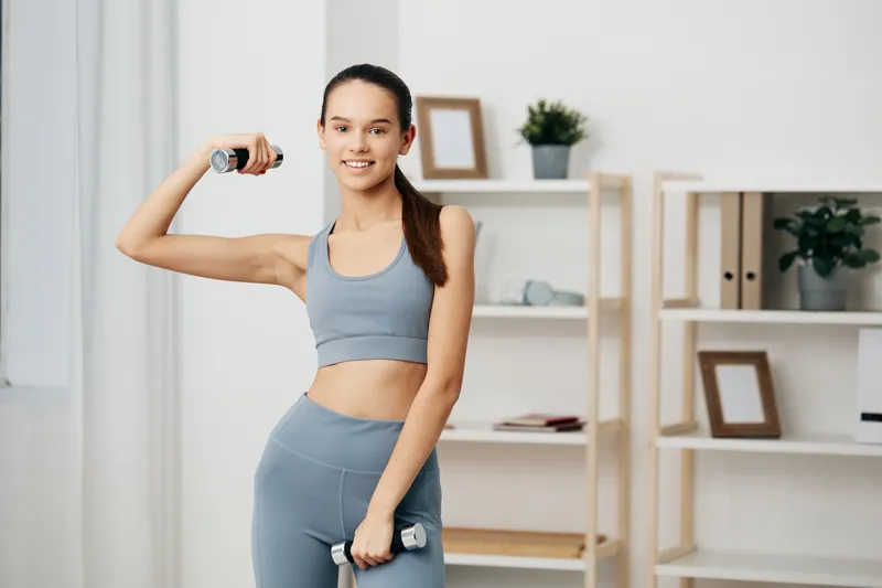 Fitness-focused woman in a blue workout set demonstrates a bicep curl with a dumbbell in a bright home gym, highlighting strength training, healthy lifestyle, and focused exercise at home.