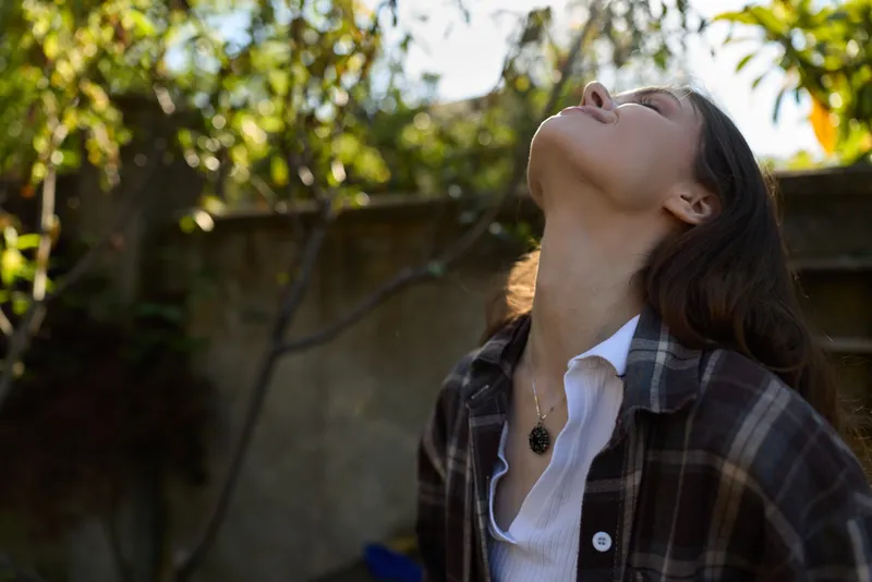 A young woman tilts her head back to catch the sun in a casual outdoor setting, conveying calm reflection and a spontaneous, authentic moment
