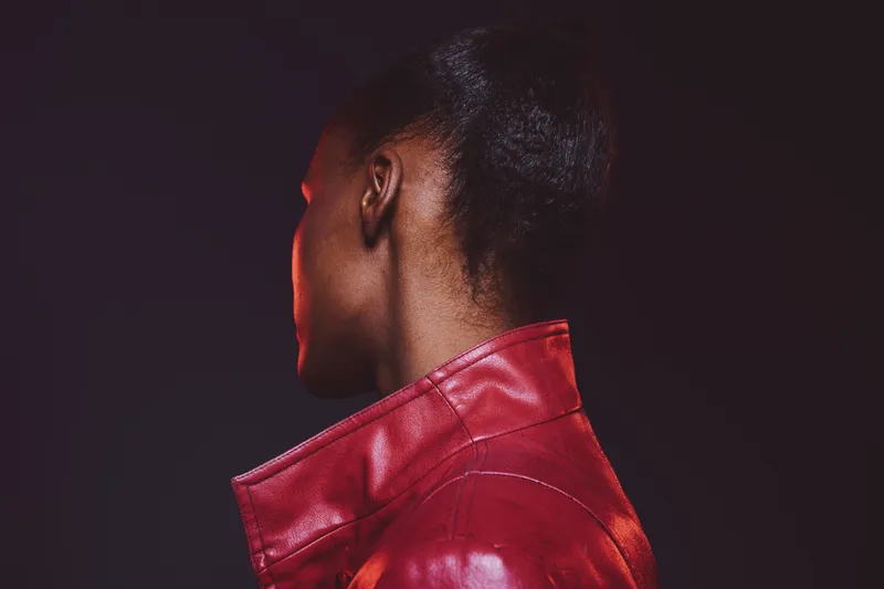 Portrait of an African woman with dark skin wearing a red leather jacket, looking away on a dark background. Concept of fashion, style, and elegance with a modern mood.