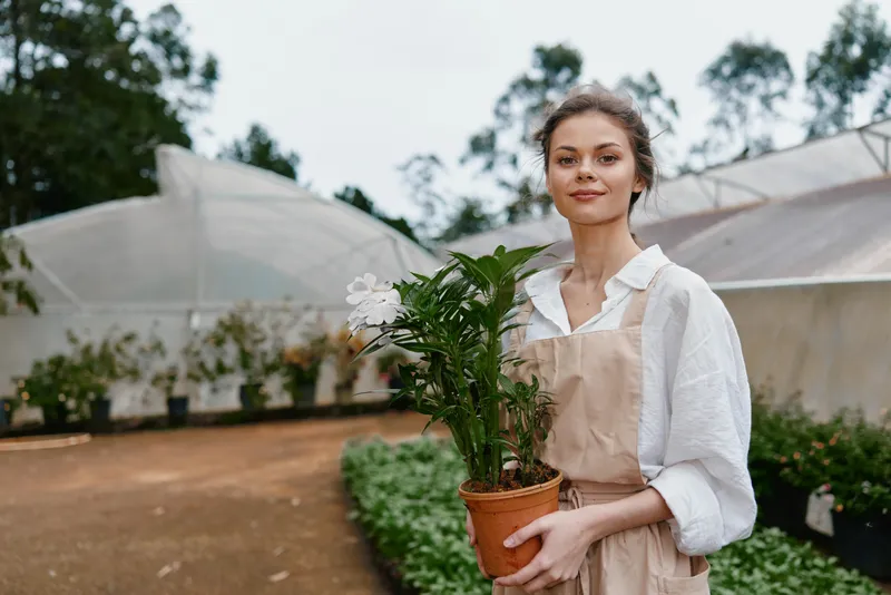 Young woman in a light apron holding a potted plant, smiling in a greenhouse filled with greenery, conveying joy and connection with nature