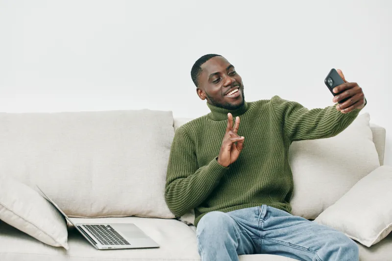 Casual portrait of a young Black man smiling and using a smartphone while sitting on a light colored sofa, conveying a sense of joy and leisure