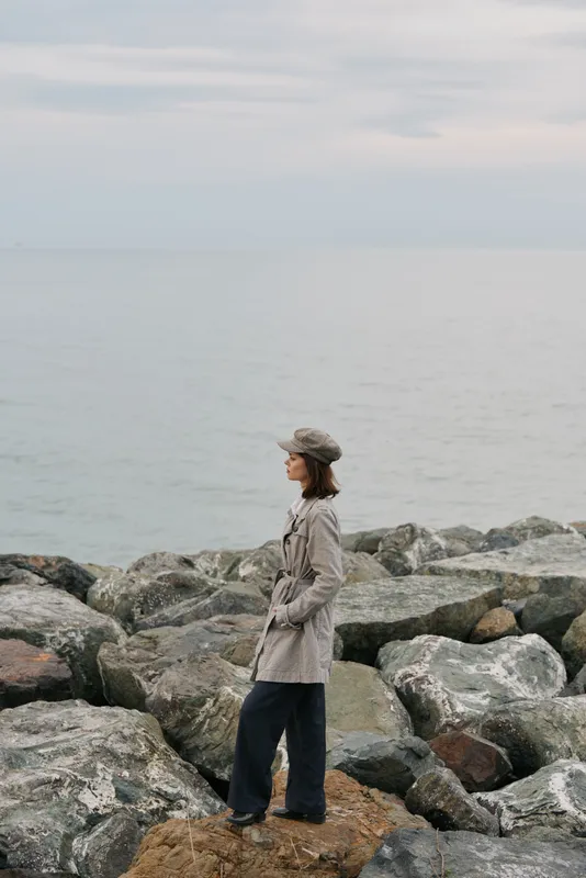 A woman in a long coat and hat stands on rocks by the sea under a cloudy sky. The calm water and overcast weather create a peaceful and contemplative mood.