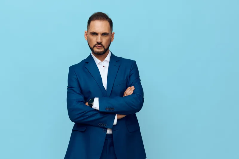A poised business professional stands against a solid blue background, projecting confidence with crossed arms and a calm, focused gaze. The isolated color backdrop emphasizes leadership.