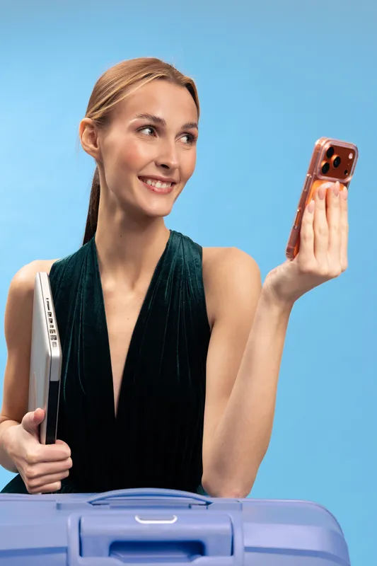 Smiling woman in dark sleeveless dress holds laptop and rose gold smartphone, standing behind blue suitcase on blue background. Concept of travel, technology, business and lifestyle.