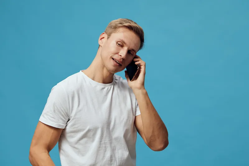 Smiling man talking on mobile phone against bright blue background. Casual white t-shirt, cheerful expression, relaxed pose, studio portrait. Communication, technology, lifestyle concept.