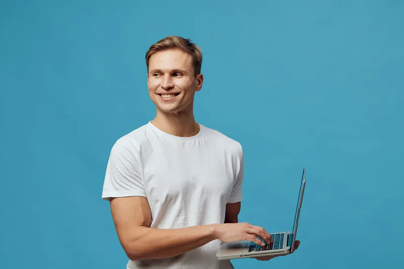 Smiling young caucasian man holding laptop wearing white t-shirt looking aside on blue background digital nomad concept remote work technology