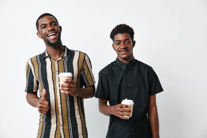 Two young black men with dark skin holding coffee cups smiling and posing on white background. Casual clothing, friendship and positive mood with beverage in hands.
