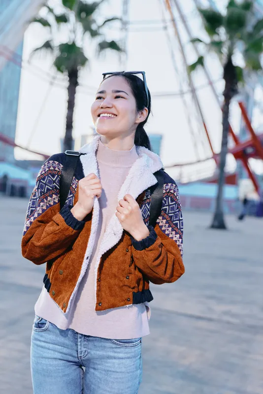 Woman smiling outdoors, wearing a patterned jacket and backpack, sunny day at a park setting, casual travel vibe, breezy style, joyful mood, street fashion for lifestyle and adventure
