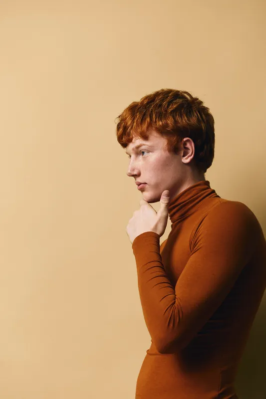 Young man with red hair wearing brown turtleneck sweater standing in profile thinking or reflecting on beige background. Side view portrait of serious male in casual clothing with hand on chin.