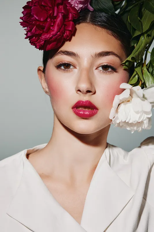 Young woman with glossy red lips and soft makeup posing with vibrant flowers in her hair, modern beauty concept including creative fashion and elegance.