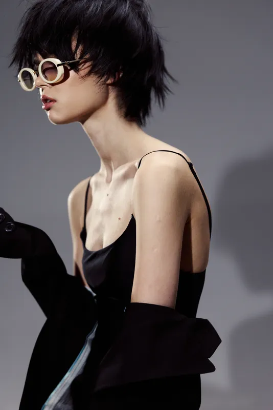 Stylish woman with short hair and round glasses wearing a black outfit in a minimalist studio setting.
