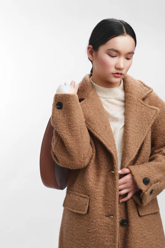 asian woman wearing a cozy brown coat adjusts her sleeve in a studio setting, conveying calm confidence and fashion consciousness with soft lighting and a minimalist backdrop.