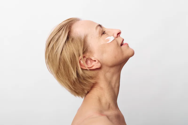 Profile of mature woman with short blonde hair applying cream on cheek, eyes closed, enjoying skincare routine. Concept of aging beauty, self-care, and natural wellness on white background.