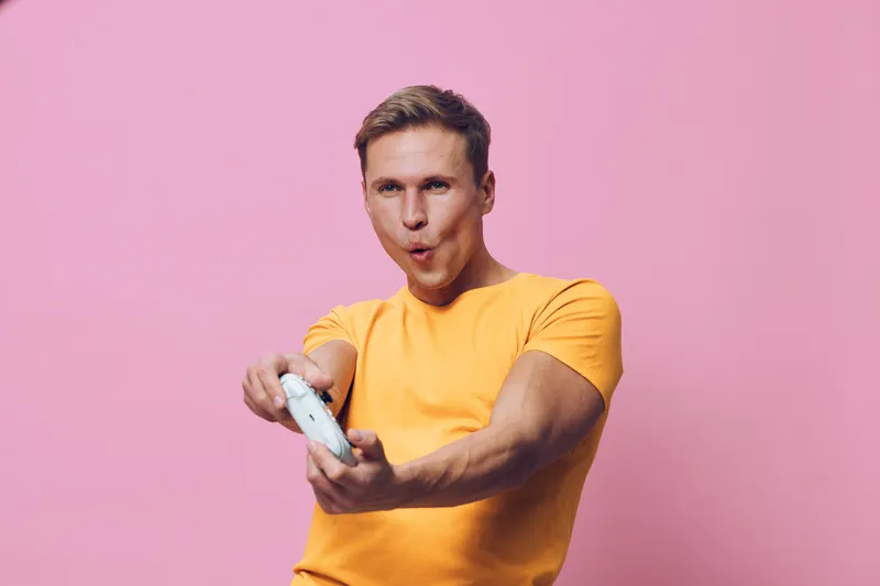 Happy young man wearing bright yellow t-shirt playing video game with controller isolated on pink background studio portrait. People lifestyle concept