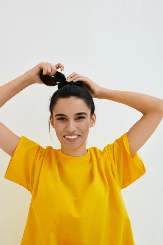 smiling woman in yellow shirt tying hair back with hands against plain white background, cheerful expression, casual style, natural lighting, youthfulness, indoor setting